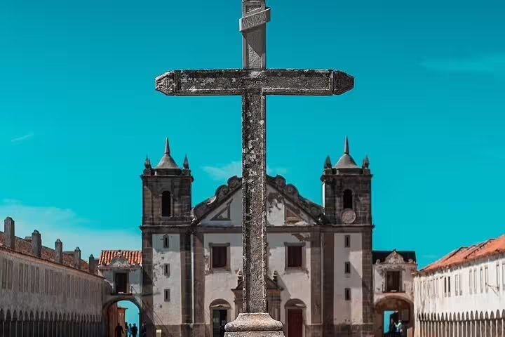 Historic stone cross with a scenic view of an ancient church in Setúbal, highlighting cultural heritage under blue skies.