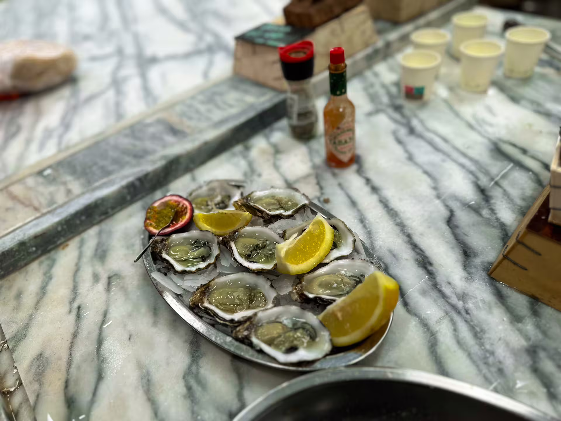 A platter of fresh oysters with lemon wedges and hot sauce on a marble counter in a Setúbal market.