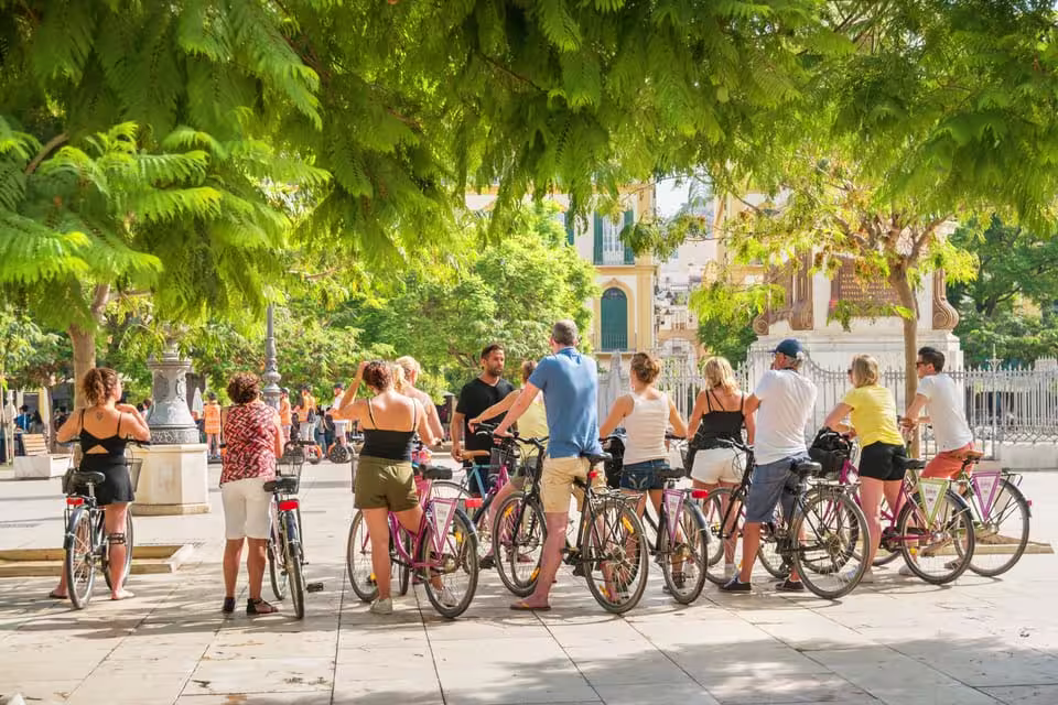 Tourists with bicycles pause under lush trees during Seville city bike tour in a vibrant plaza.