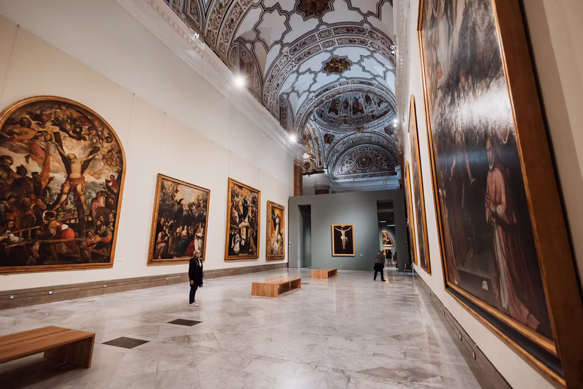 Visitors admire historic paintings in the grand hall of Seville's Fine Arts Museum, showcasing exquisite Spanish art and architecture.