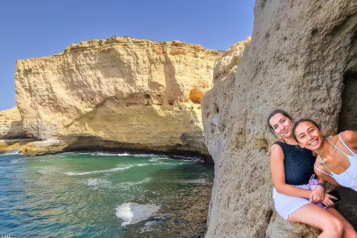 Two smiling women enjoying scenic cliffs and turquoise waters on the Algarve coast during a Seville to Lisbon transfer tour.