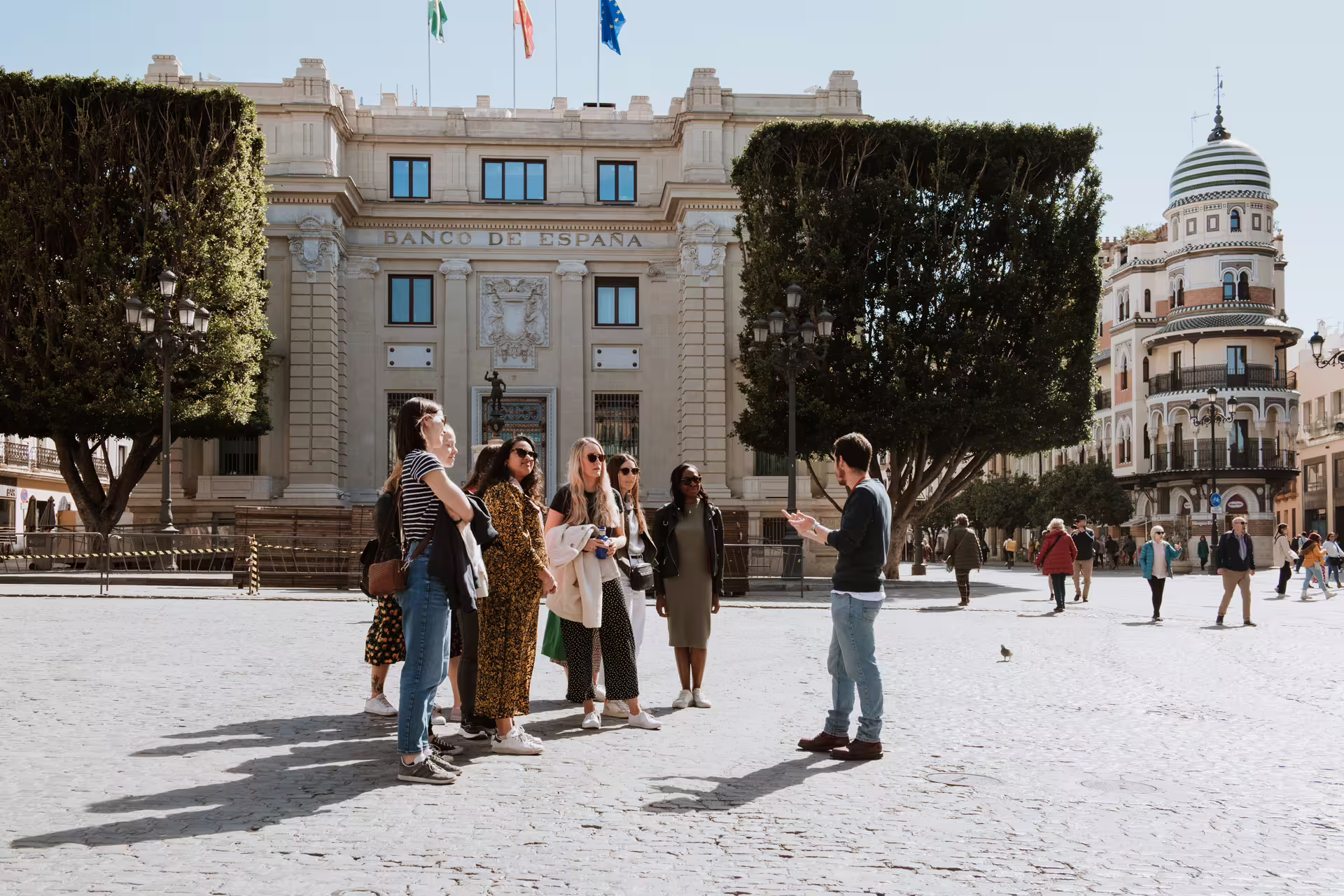 A guide leads a private food tour in Seville's historic plaza, with tourists exploring local culinary delights and architecture.