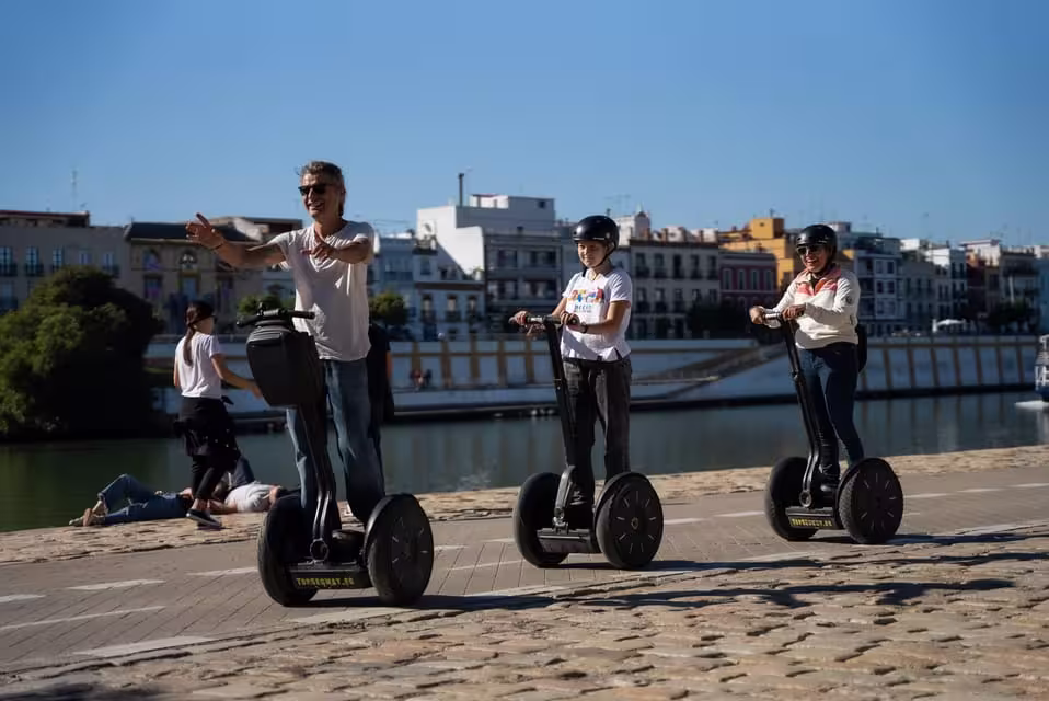 Tourists on Segways along the riverside promenade in Seville, with cityscape in the background.