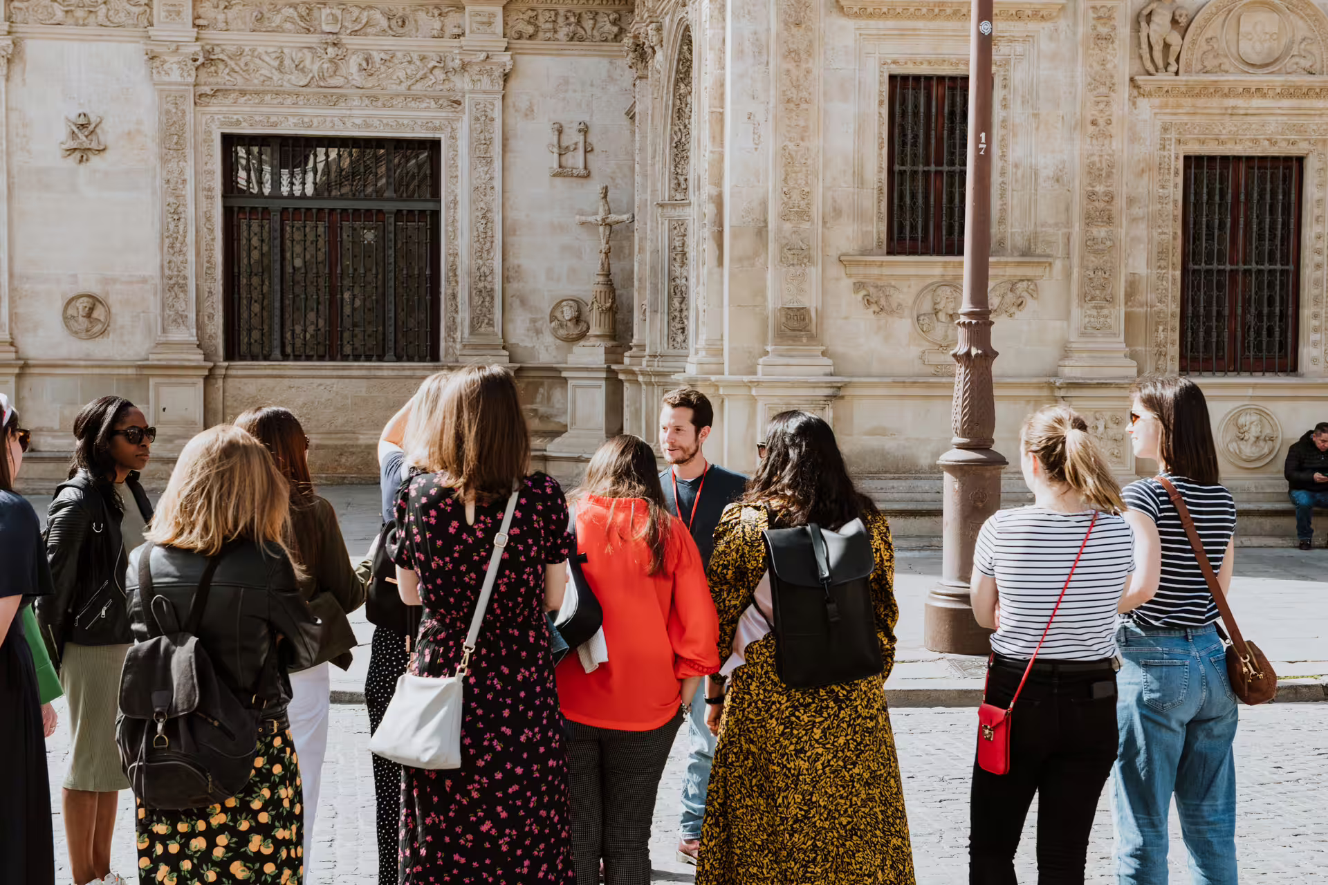 A small group of tourists listens to a guide during a Seville walking tour, exploring historic architecture in a sunny plaza.