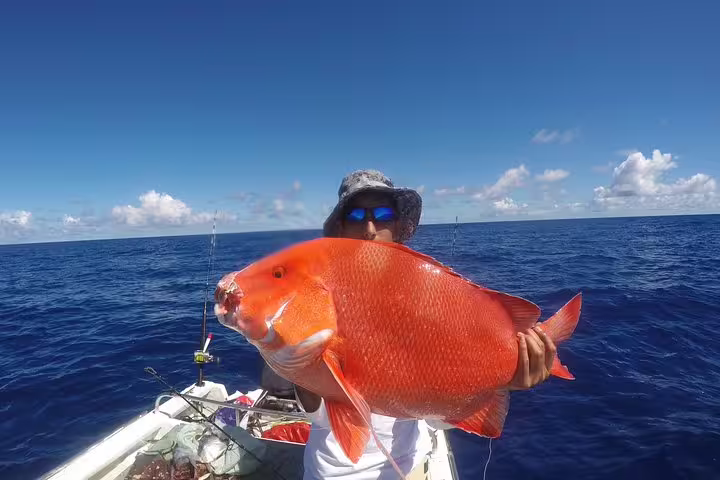 A fisherman proudly holding a large, vibrant red fish aboard a boat under a clear blue sky in Seychelles.