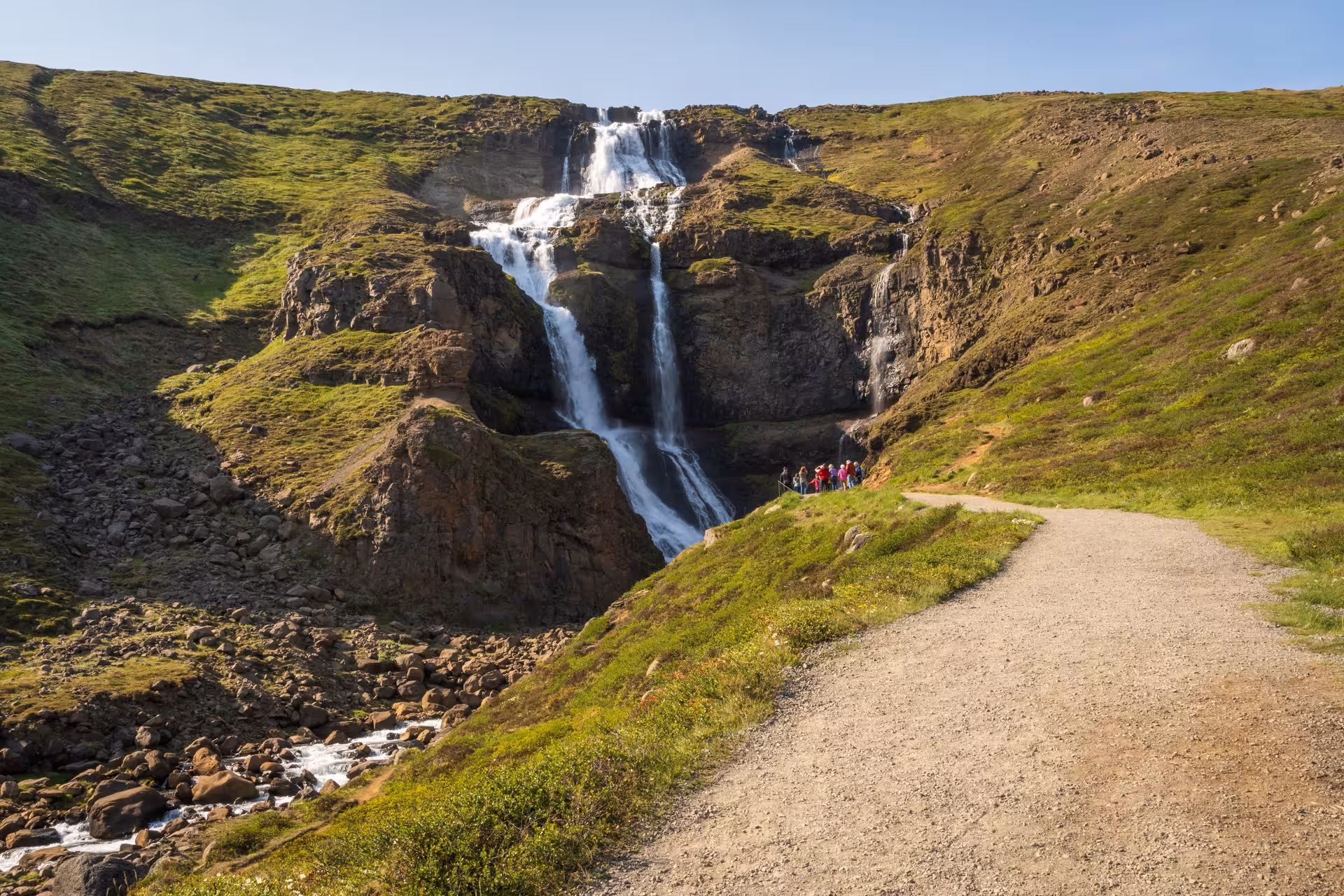 Multi-tiered waterfall cascading down green rocky cliffs with hikers on a gravel path in Iceland