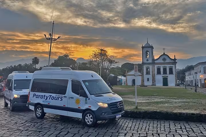 Shuttle vans parked at sunset in front of a historic church, part of a shared transfer service to Angra dos Reis.