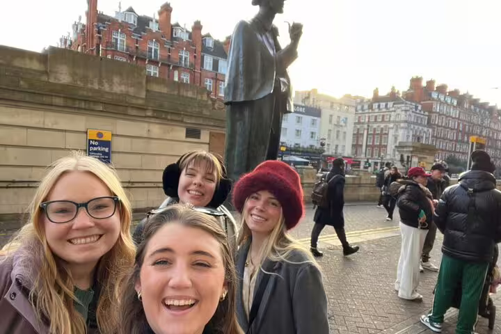Four smiling women posing for a selfie in front of a bronze statue on a London street