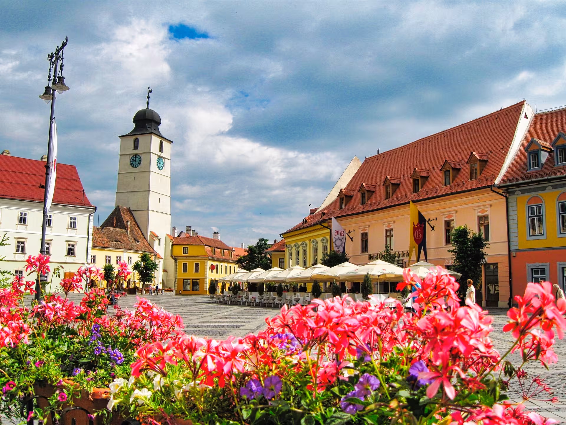 Sibiu Grand Square with Council Tower, colorful buildings, outdoor café umbrellas, and pink flowers in foreground