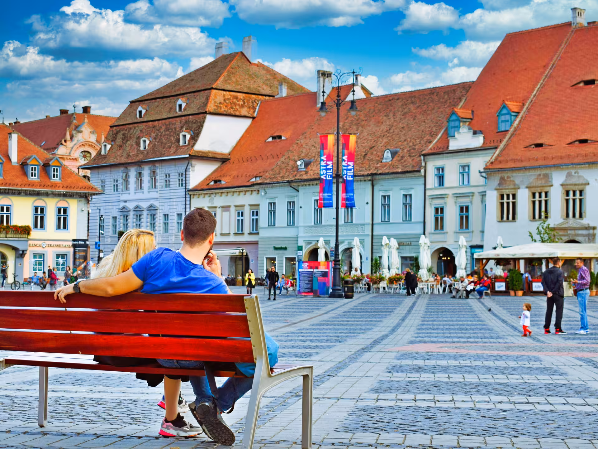 Couple sitting on a bench in Sibiu's Grand Square with colorful historic buildings and Astra Film banners