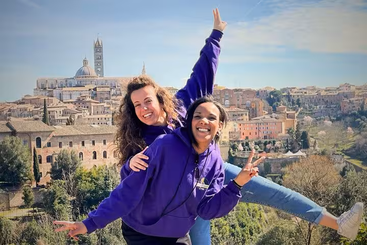 Two smiling women in purple shirts posing with Siena's historic skyline in the background on a sunny day.