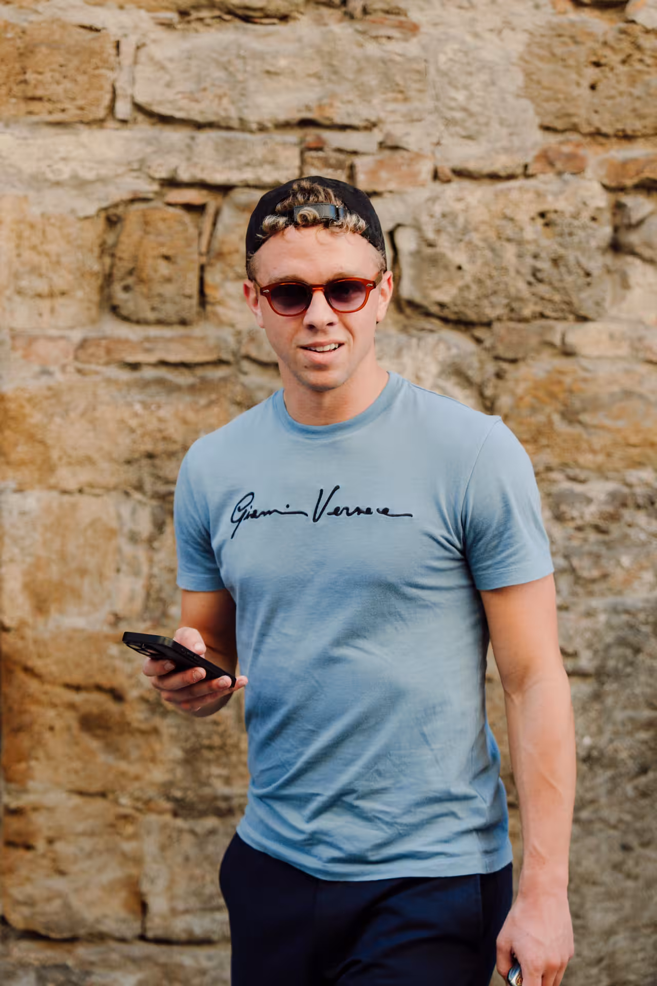 Young man in Gianni Versace t-shirt and sunglasses holding smartphone against stone wall in Siena
