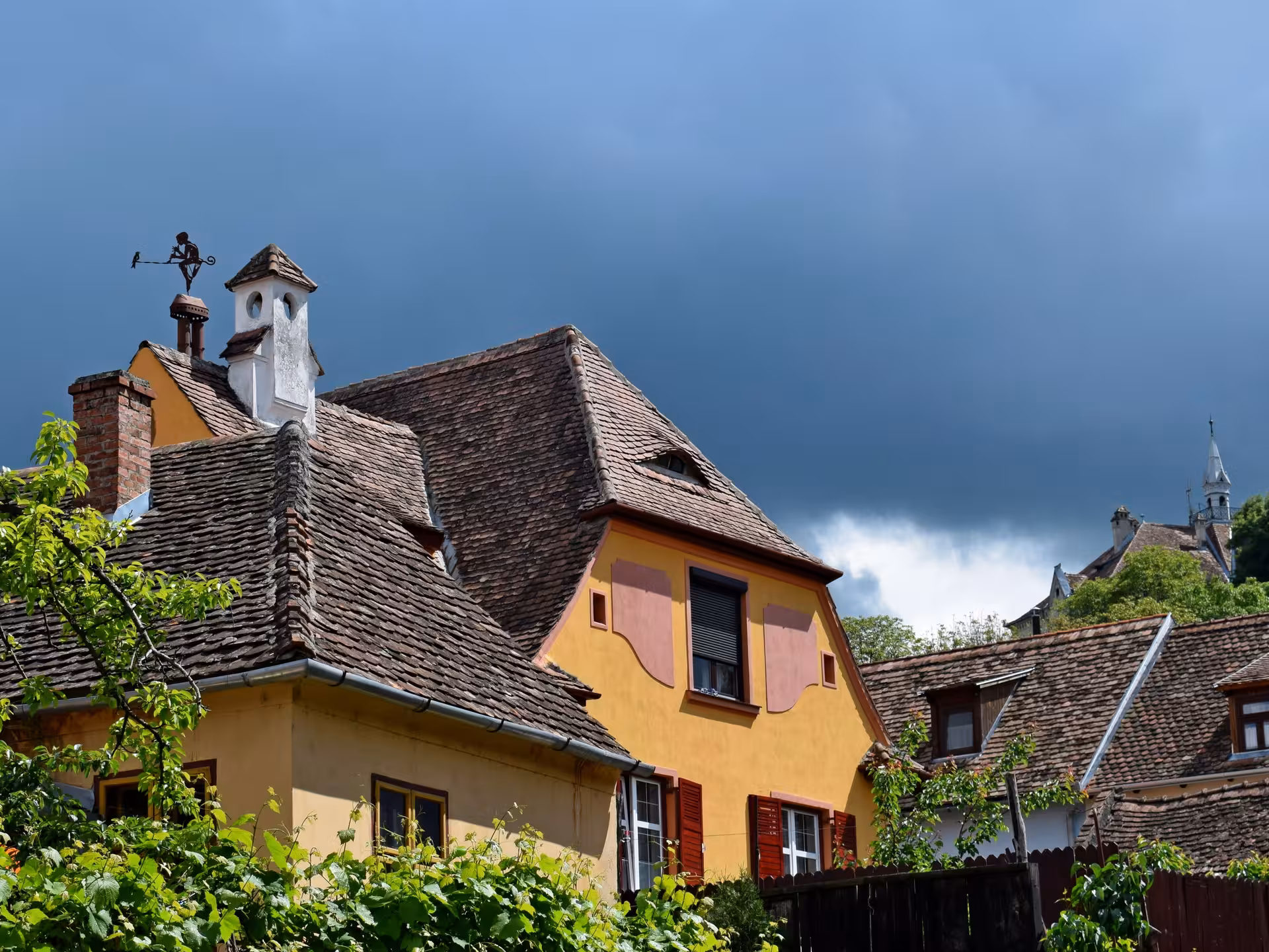 Historic yellow house with tiled roof and weather vane under dramatic stormy sky in Sighisoara