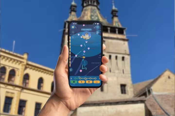 Hand holding smartphone with Sighisoara self-guided tour app map in front of the Saxon Citadel clock tower