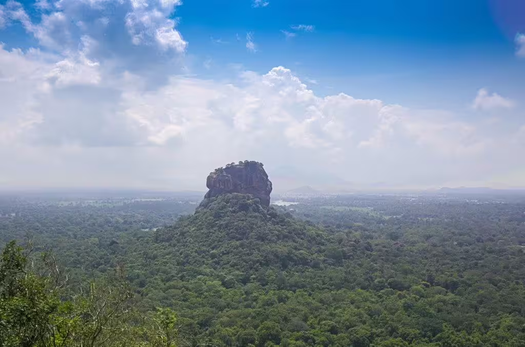 Aerial view of Sigiriya Rock Fortress rising majestically from lush green forests under clear blue skies in Sri Lanka.