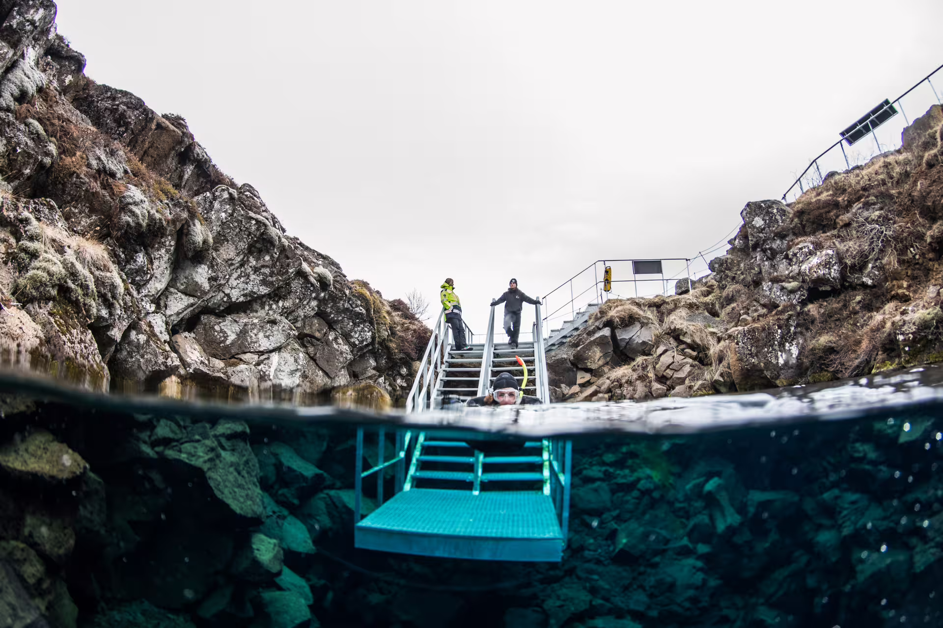 Snorkeler in crystal clear Silfra fissure water with metal entry stairs and rocky canyon walls in Iceland