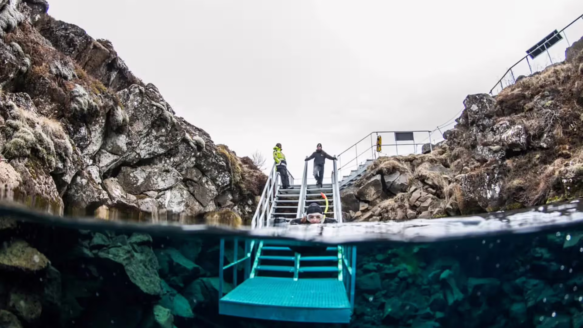 Snorkeler entering crystal clear Silfra fissure water via metal stairs with rocky canyon walls in Iceland