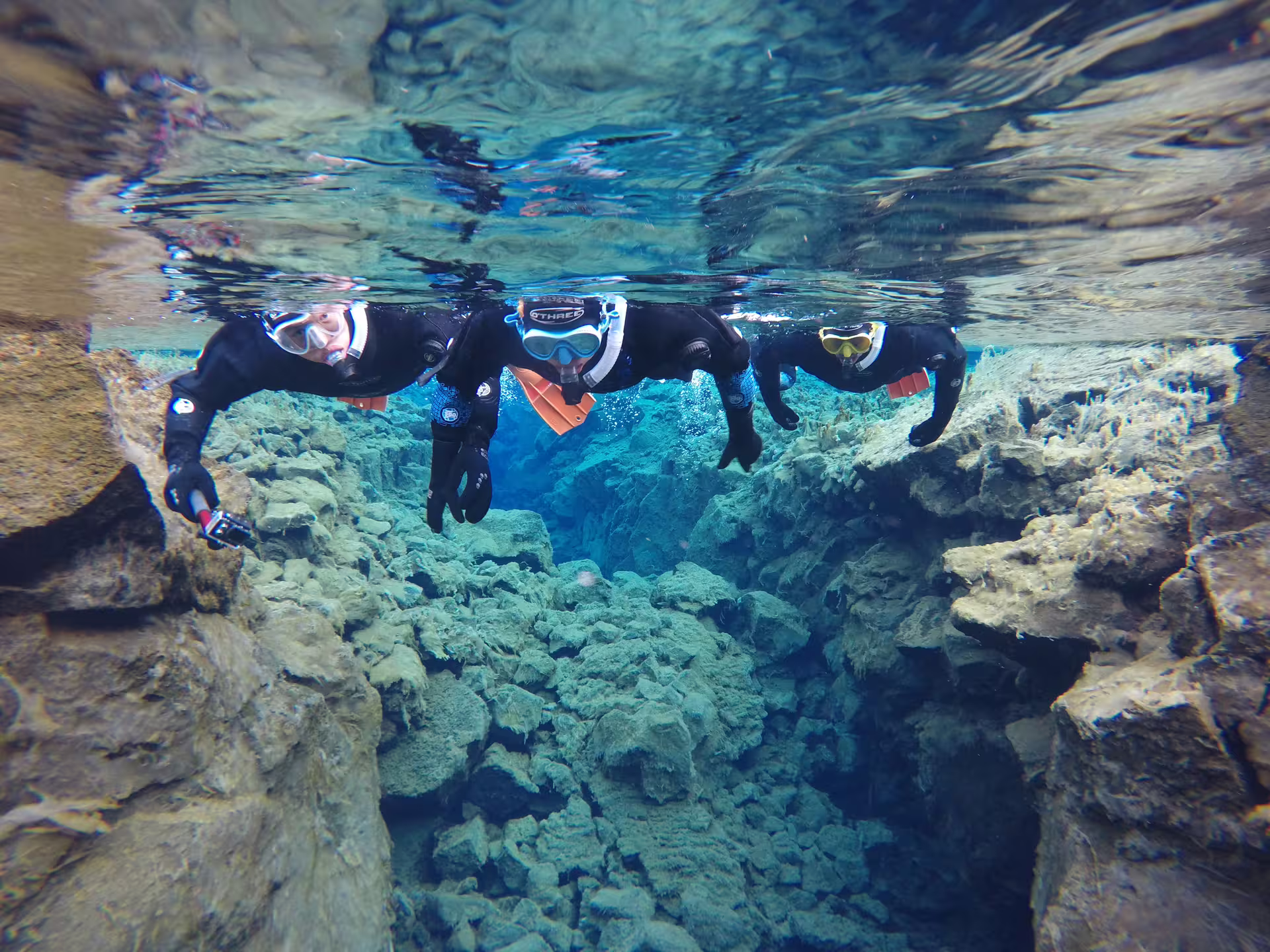 Three snorkelers in wetsuits floating above rocky fissure in crystal clear Silfra waters, Thingvellir Iceland