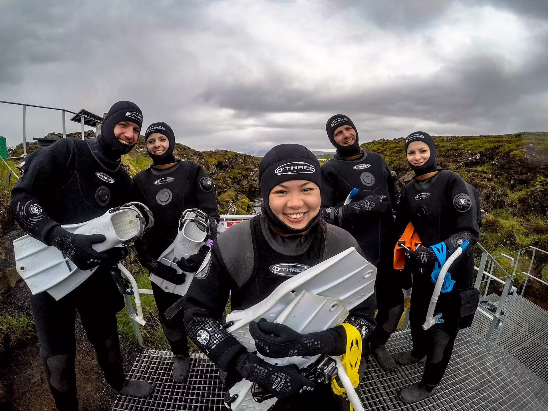 Five smiling divers in O'Three drysuits holding fins at Silfra fissure, Iceland