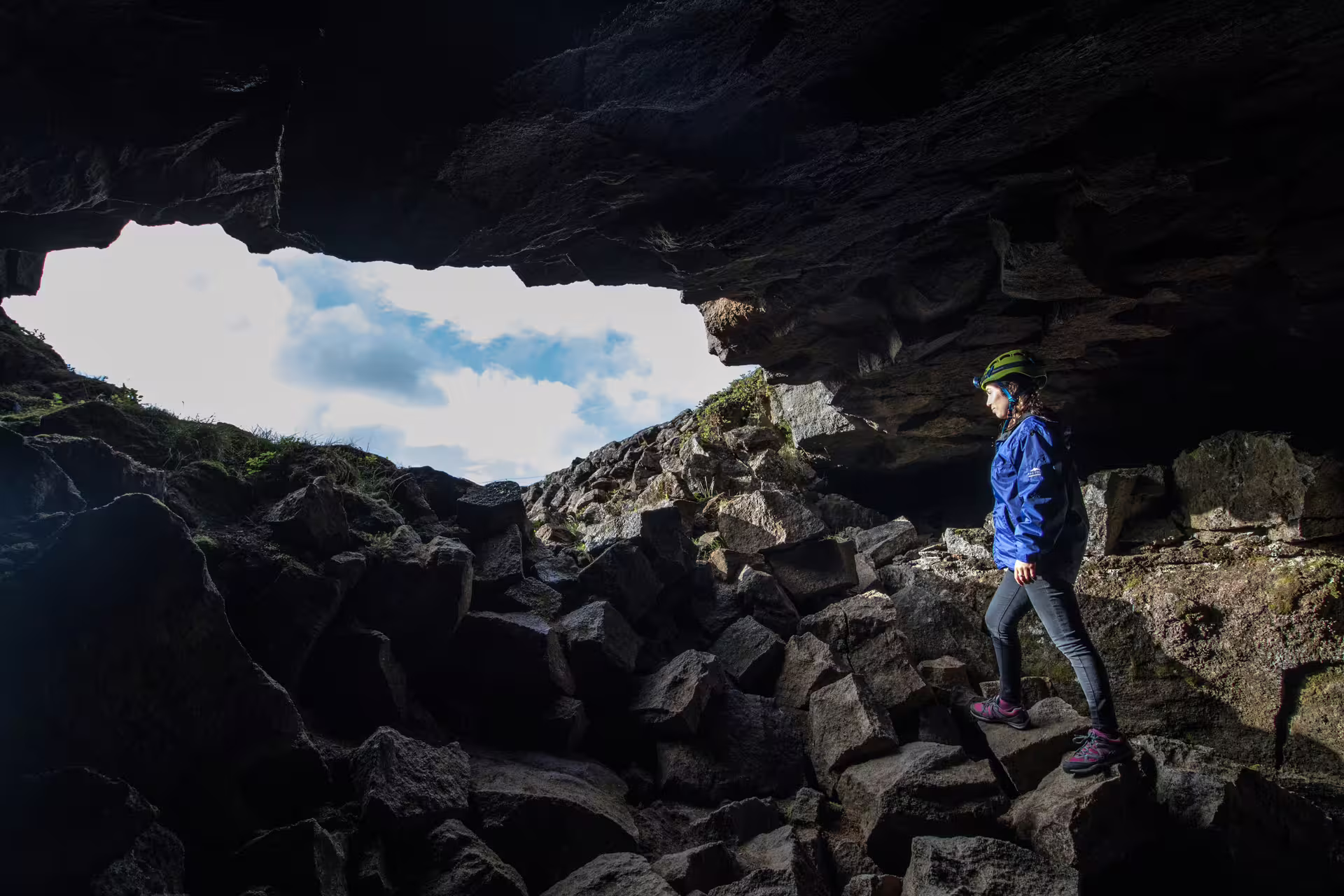 Explorer in protective gear navigates rocky terrain inside Leidarendi Lava Tunnel, Iceland, with daylight streaming through the entrance.