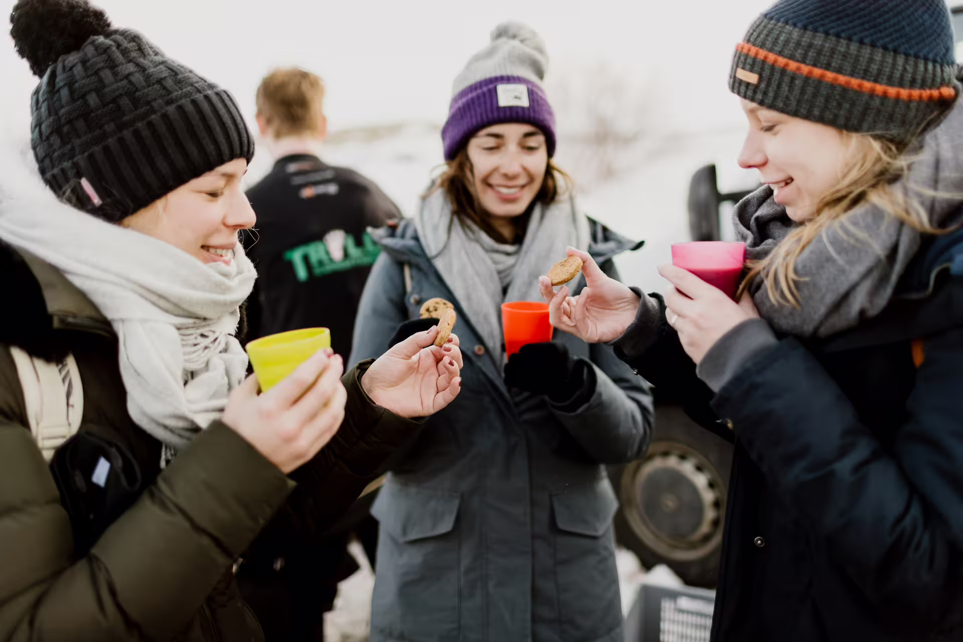 Three women in winter gear sharing cookies and hot drinks outdoors in snowy Thingvellir Iceland