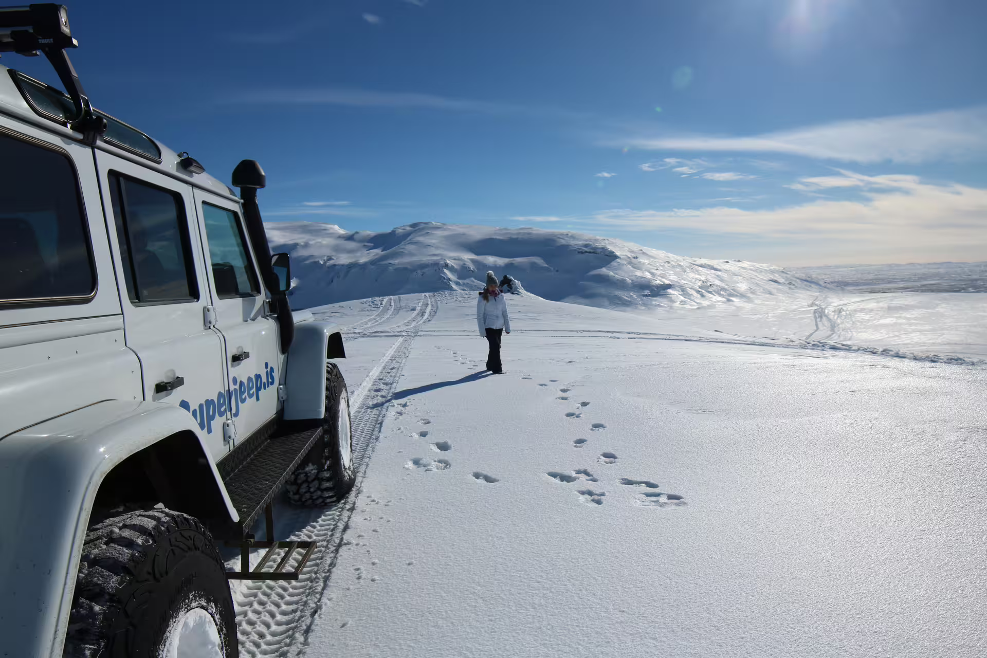 A Superjeep parked on snowy terrain with a person walking towards Iceland's stunning Golden Circle under a clear blue sky.