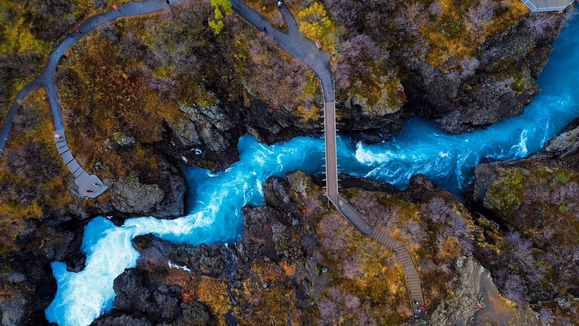 Aerial view of turquoise glacial river rushing through lava rock canyon with footbridge in West Iceland