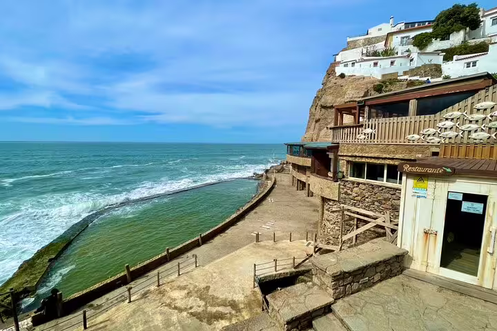Scenic coastal view in Sintra with cliffside buildings and ocean waves, highlighting the allure of the Sintra & Cascais tour.