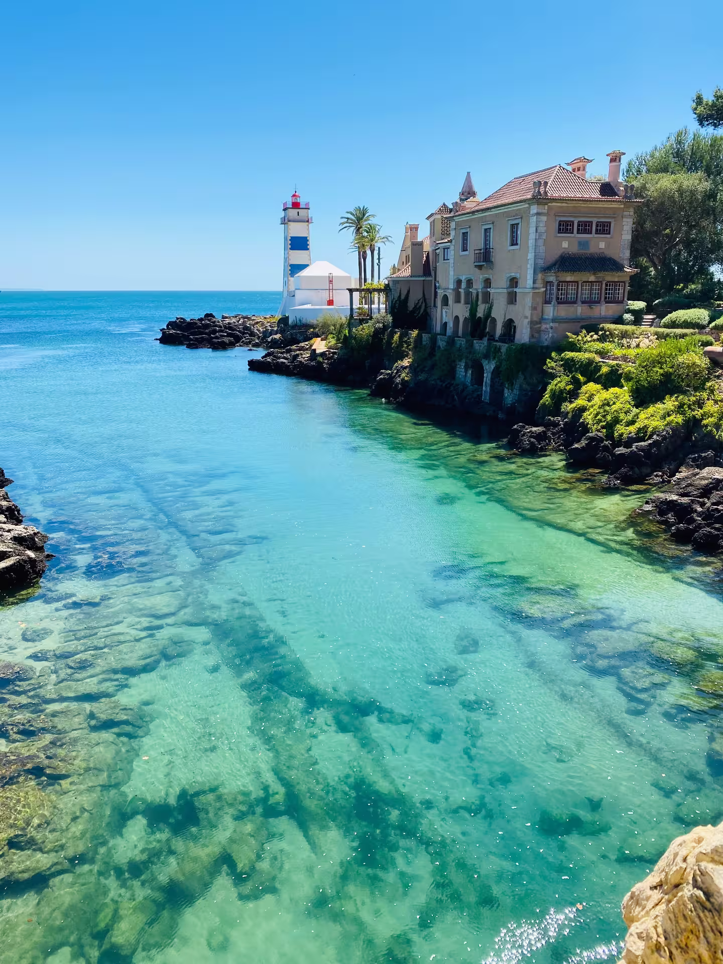 Scenic view of a coastal villa and lighthouse in Cascais with crystal-clear turquoise waters under a bright blue sky.