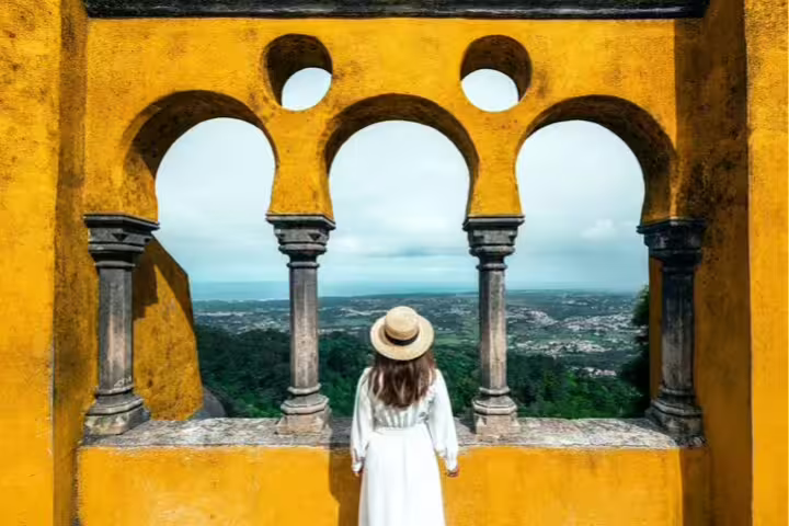 Woman in a white dress and straw hat gazes through vibrant arches at panoramic Sintra landscape on a private tour with wine tasting.