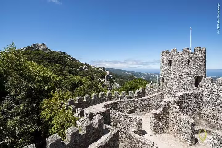 View of the historic stone walls and lush landscape of Sintra's Moorish Castle on a sunny day, a highlight of the Sintra, Pena Palace, and Cascais full-day tour from Lisbon.