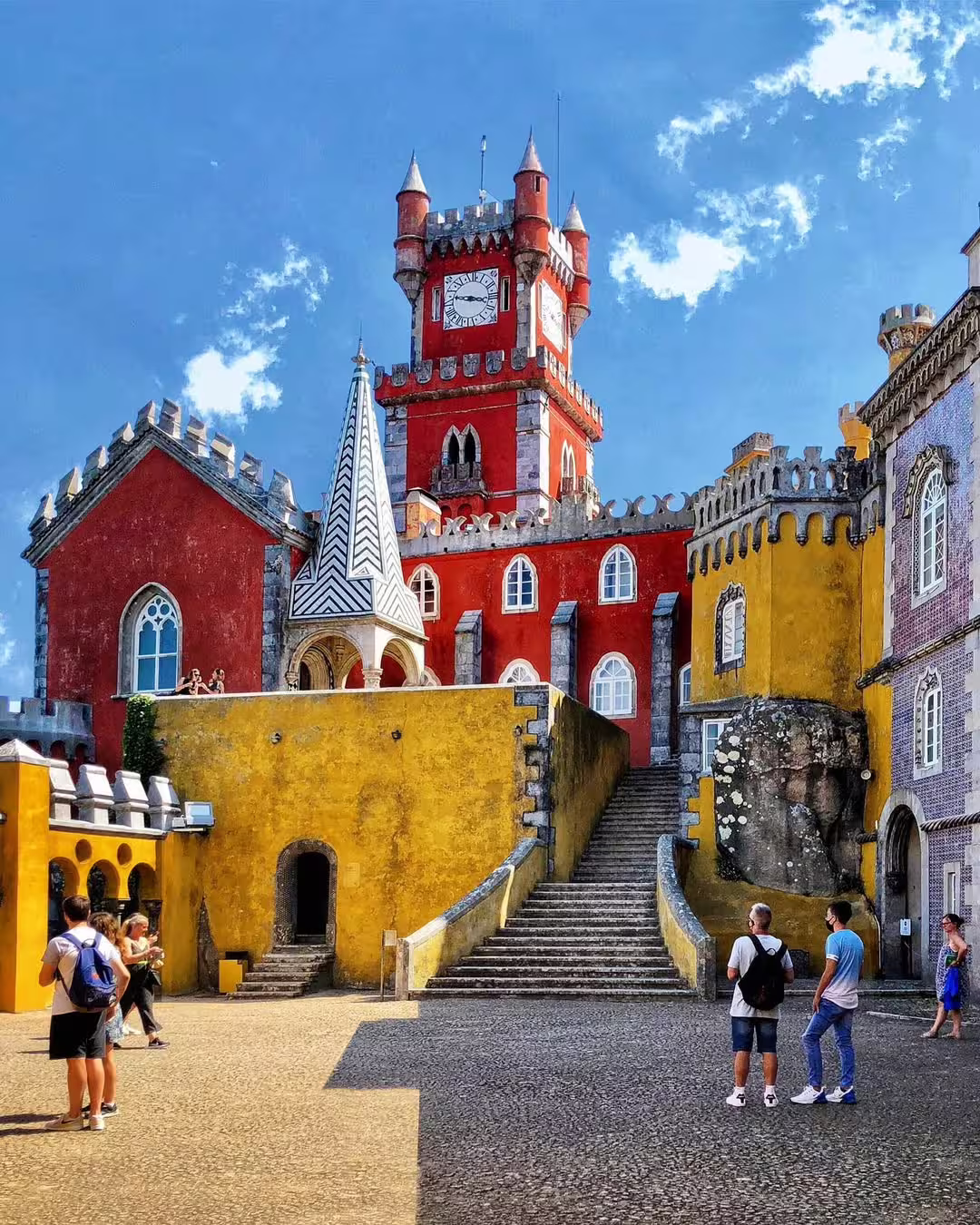Vibrant view of Sintra's colorful Pena Palace with tourists exploring the historic architecture on a sunny day.