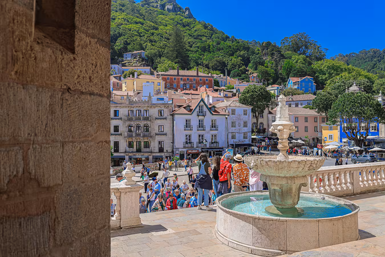 A vibrant scene in Sintra's town square with a historic fountain and colorful buildings surrounded by lush hills.