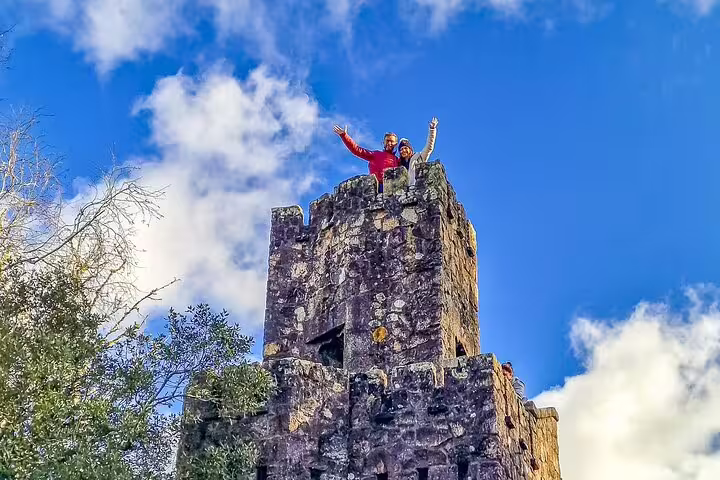 Two tourists wave from atop a historic stone tower in Sintra, Portugal, under a vibrant blue sky, during a private tour.