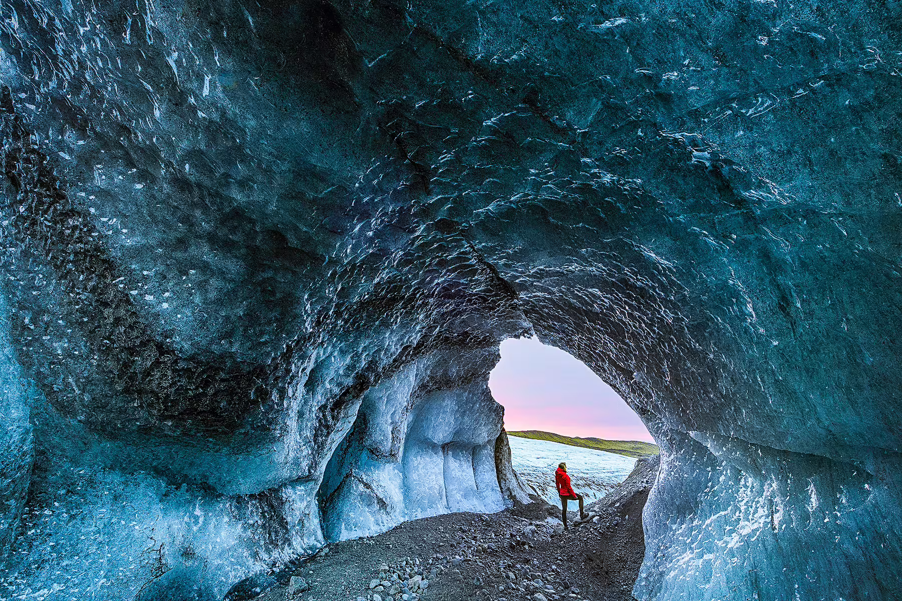 Explorer in red jacket standing at the entrance of a blue ice cave in Skaftafell glacier at dusk