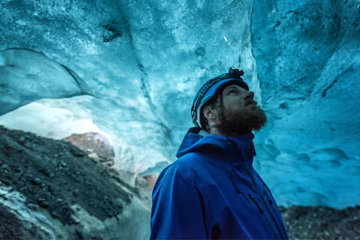 Bearded explorer in blue jacket and helmet gazing up at glowing turquoise ice cave walls at Skaftafell glacier
