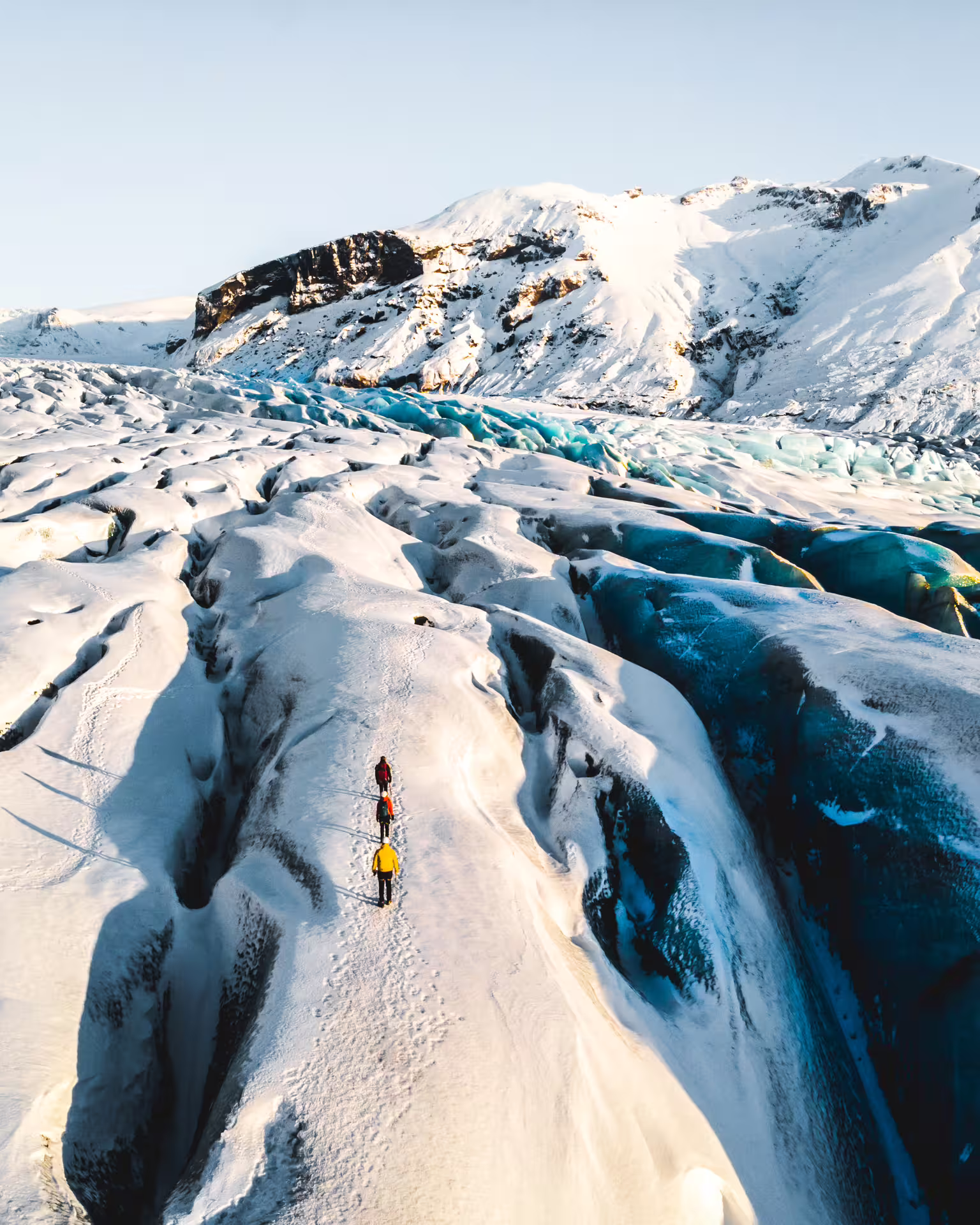 Three hikers trekking across Skaftafell glacier with blue ice crevasses and snow-covered mountains in Iceland