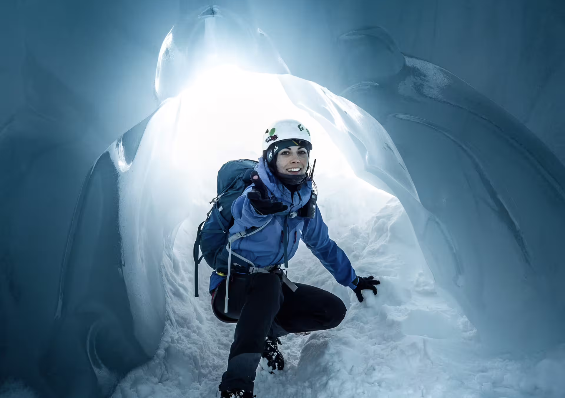 Smiling female hiker in blue jacket and white helmet crawling through a glacial ice cave at Skaftafell