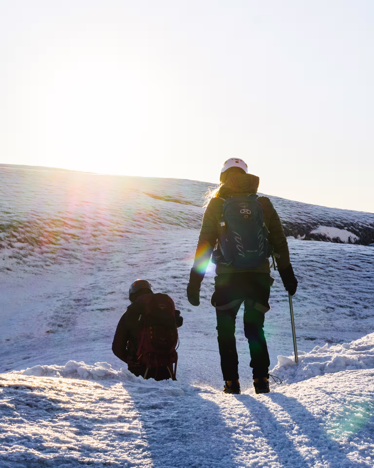 Two hikers with backpacks and helmets ascending a snowy glacier slope in bright winter sunlight