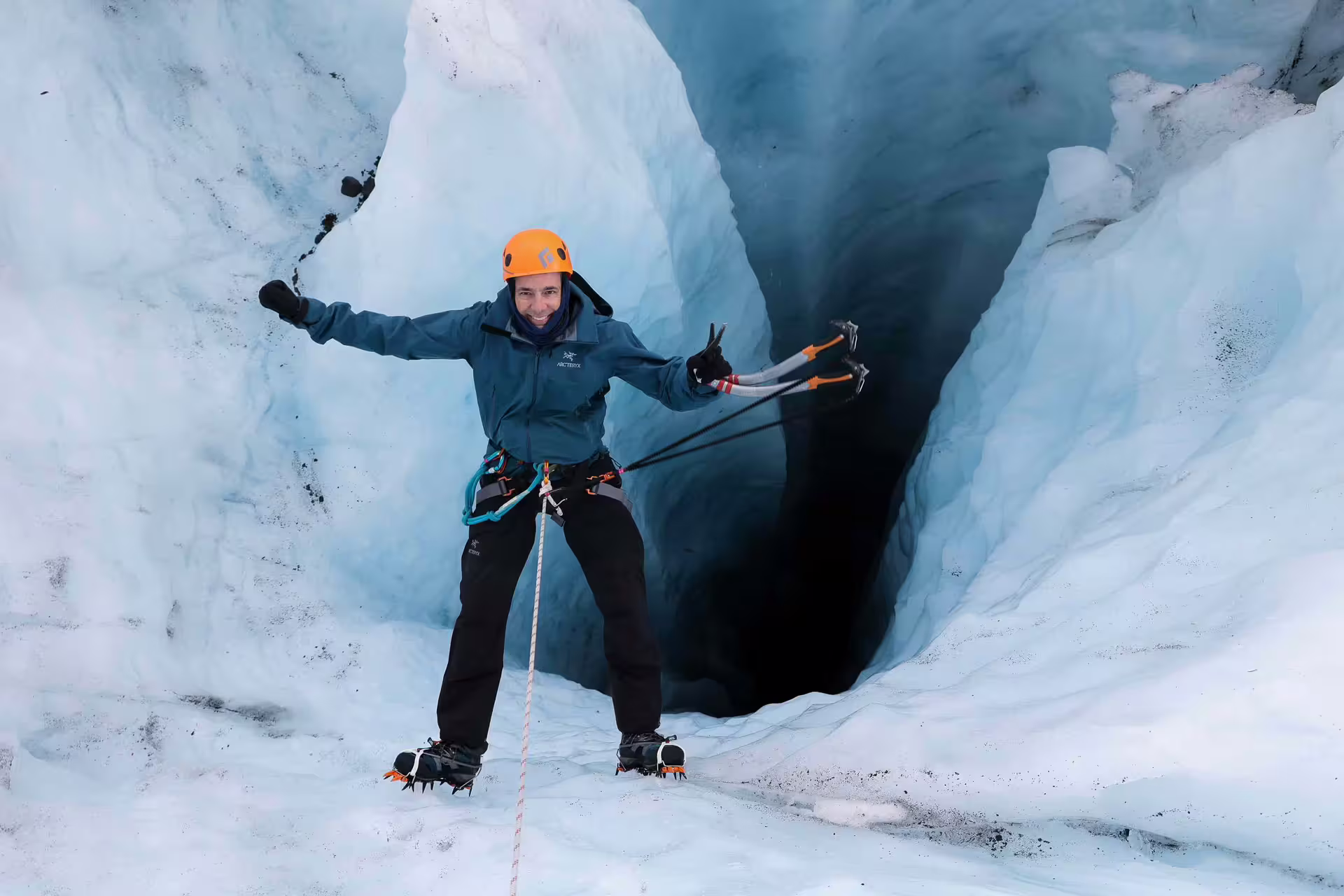 A smiling adventurer in full gear poses with ice axes at a glacial cave entrance on a Skaftafell ice climbing tour in Iceland.