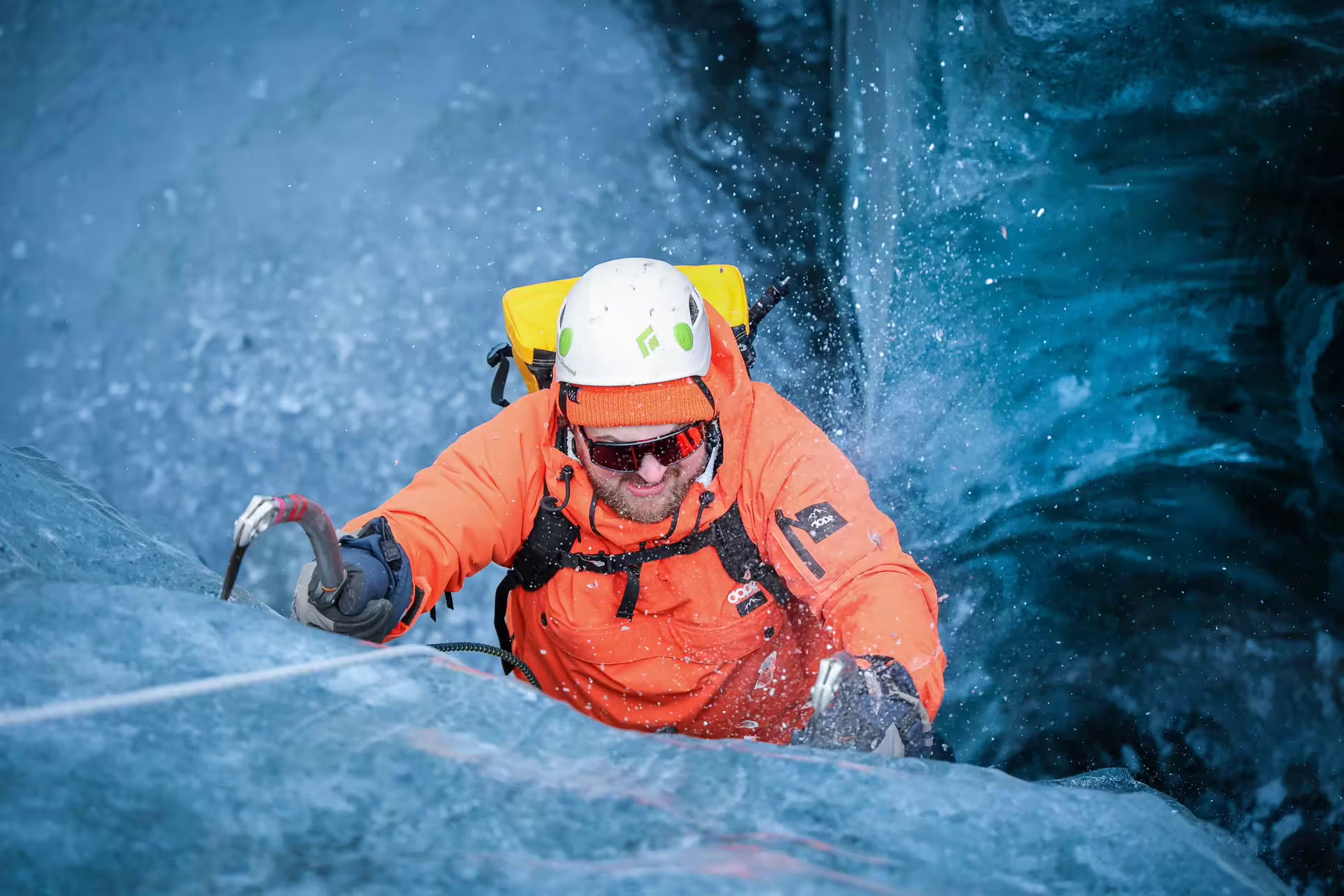 A climber in orange gear scales a blue glacial ice wall with an ice axe on the Skaftafell glacier tour