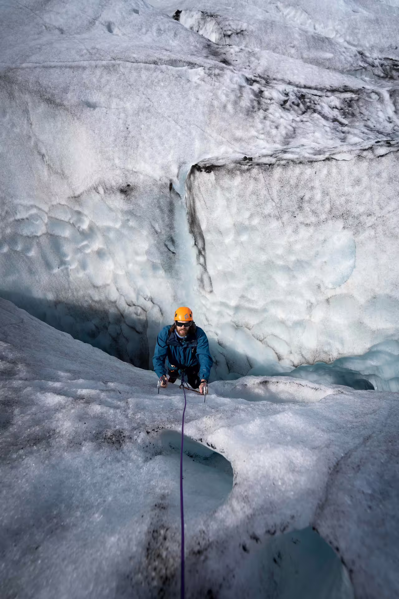 A guided climber in an orange helmet ascends a steep blue ice wall on the Skaftafell Glacier ice climbing tour in Iceland