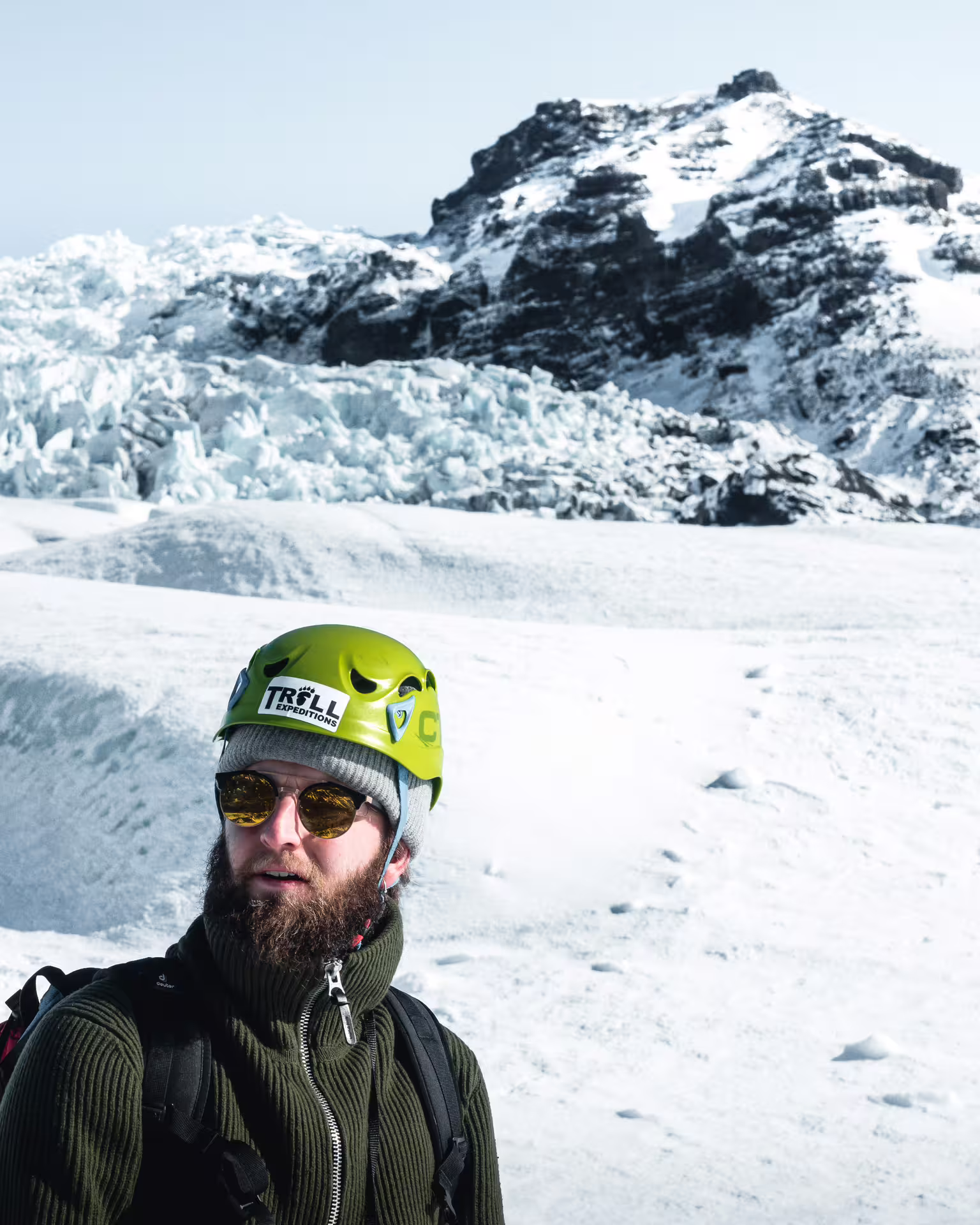 Bearded hiker wearing green Troll Expeditions helmet and sunglasses on Skaftafell glacier with snowy mountain backdrop