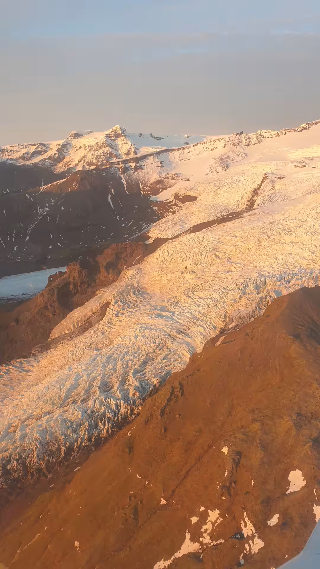 Aerial view of a glacier flowing over rugged volcanic mountains bathed in golden sunset light, Skaftafell Iceland