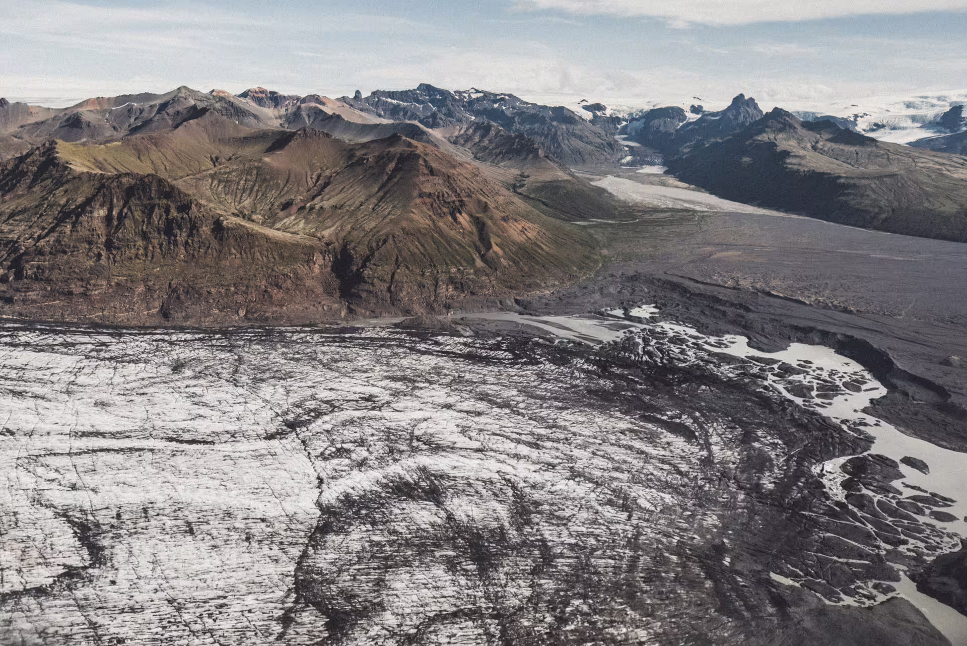 Aerial view of a dark ash-covered glacier with crevasses and rugged volcanic mountains in Skaftafell, Iceland