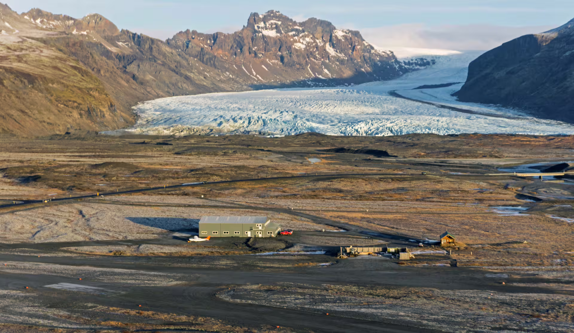 Aerial view of Skaftafell glacier tongue flowing between mountains over black sand plains with a small lodge below