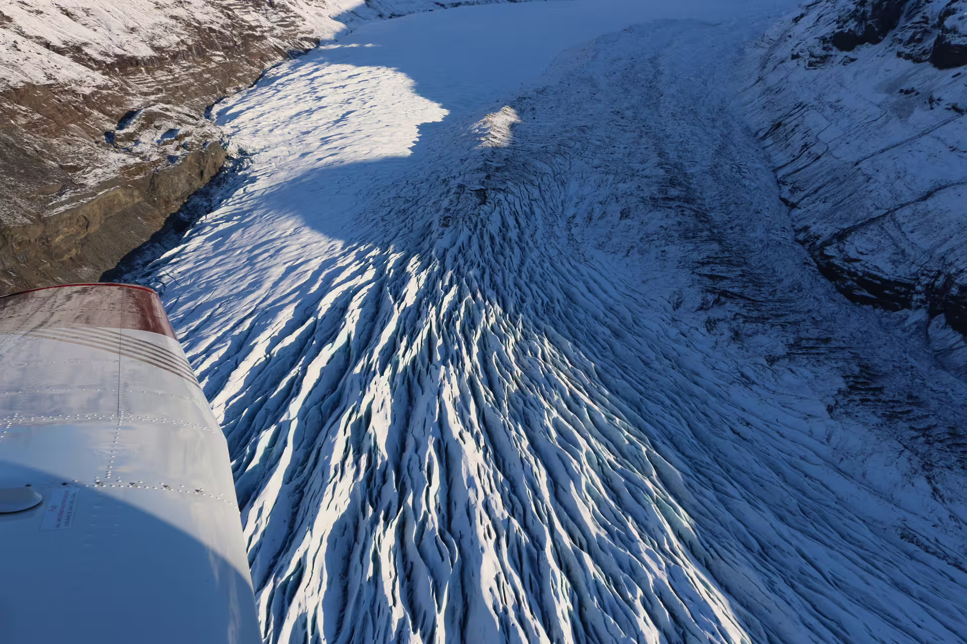 Aerial view of heavily crevassed glacier flowing through rocky mountain valley, shot from small plane wing