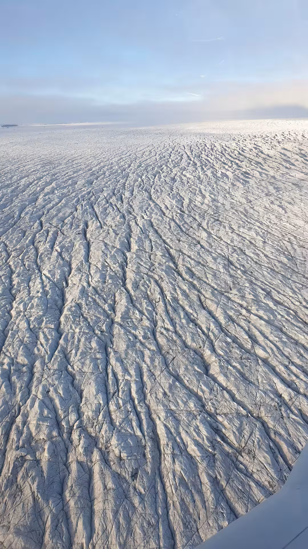 Aerial view of a vast glacier with rippled ice textures and crevasses under a pale blue sky