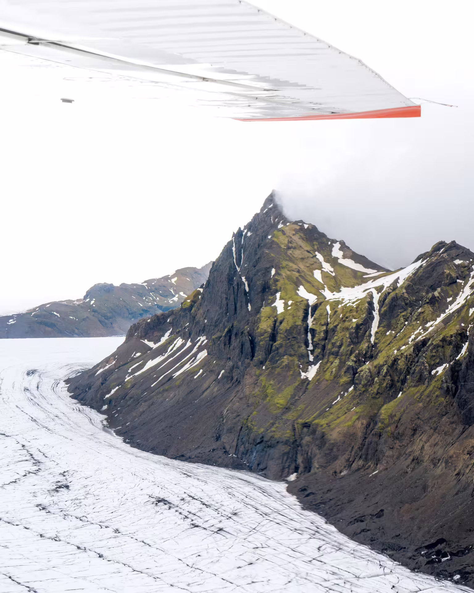 Aerial view of Skaftafell glacier flowing beside rugged snow-patched mountain peaks from small plane window