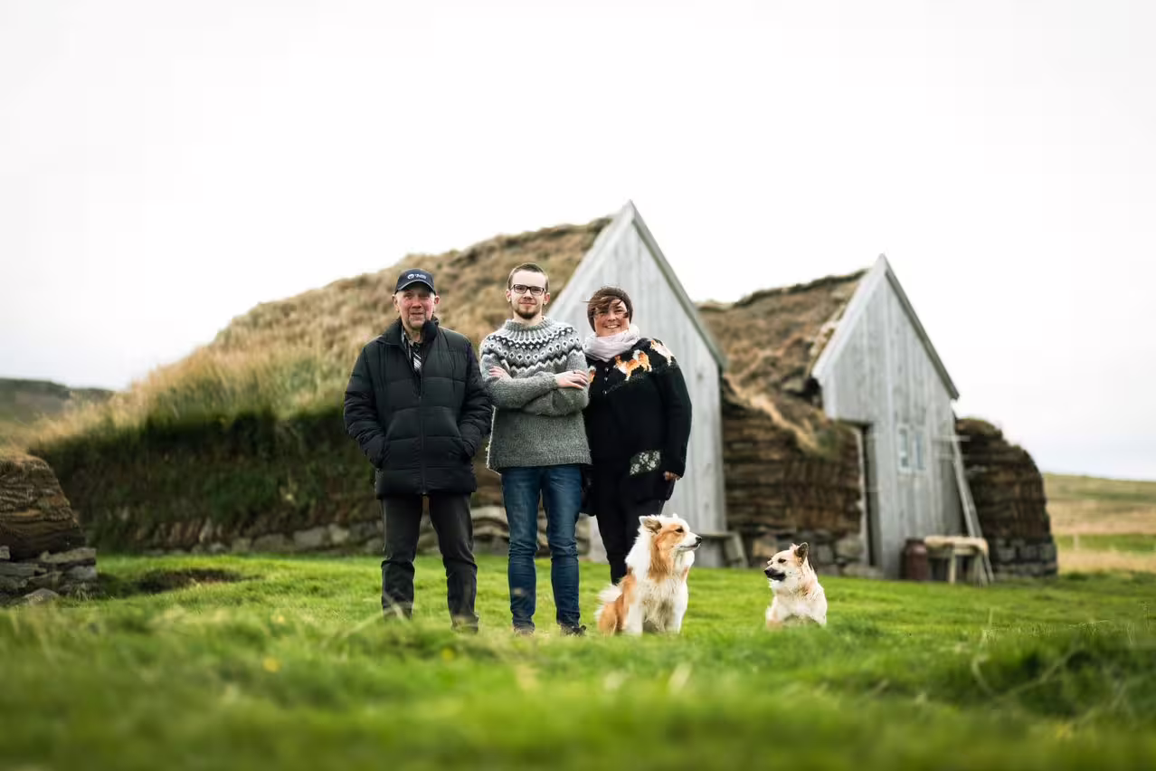 Three people and two dogs standing in front of traditional turf houses in Skagafjordur, Iceland